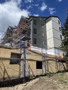 The south east side of the building has vertical siding and grey shingle siding. The new windows are installed. The garage walls are under construction and a worker is seen on the scaffolding in front of the wall.